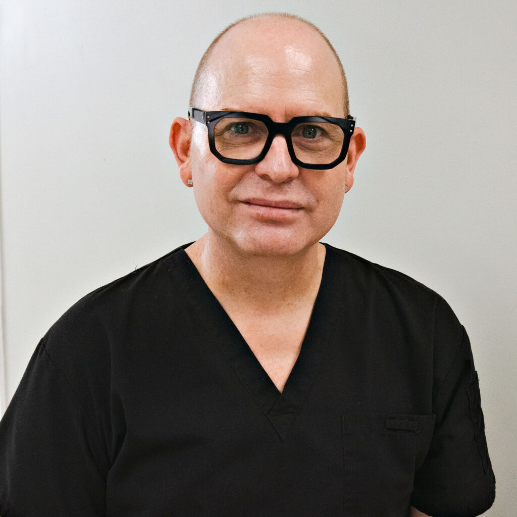 Person in black scrubs wearing glasses against a plain background