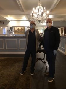 Two healthcare workers in masks standing in a well-lit room with a chandelier