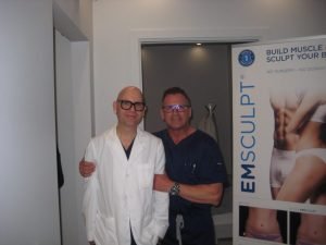 Two men stand near an Emsculpt advertisement at a clinic